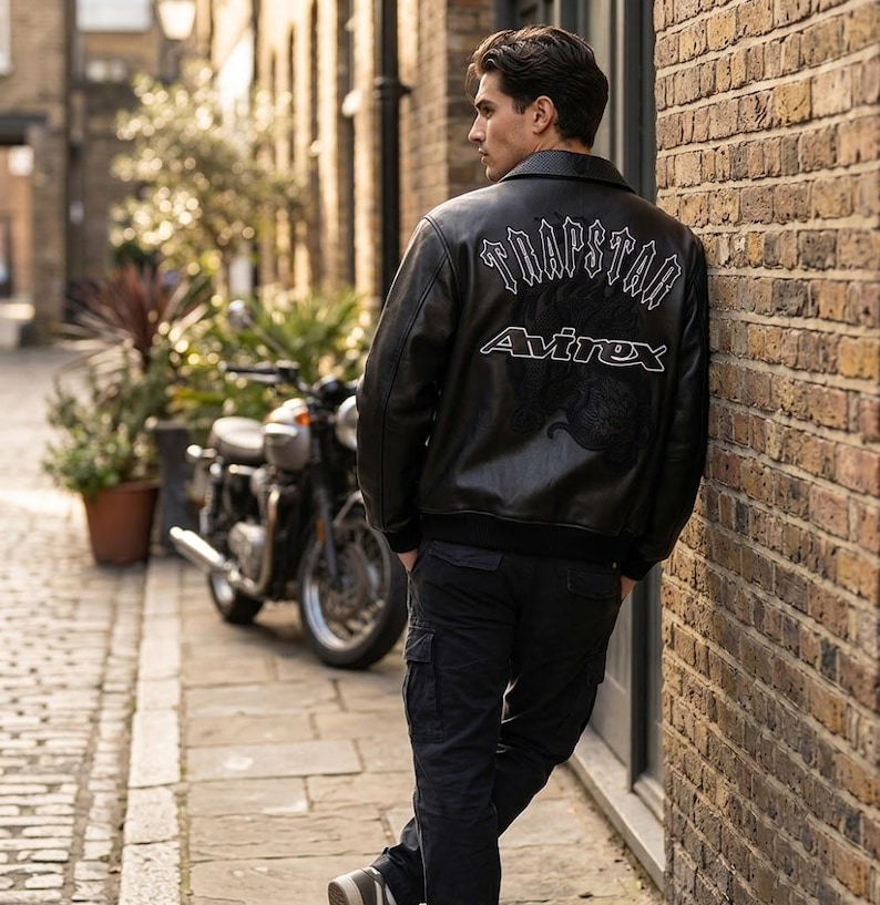 K&ouml;nnte beinhalten: Schwarze Lederjacke mit dem Text "TRAPSTAR" und "Avirex" in Wei&szlig;. Die Jacke hat einen klassischen Bomberstil mit geripptem Kragen und B&uuml;ndchen. Die Person lehnt an einer Backsteinmauer neben einem Motorrad.