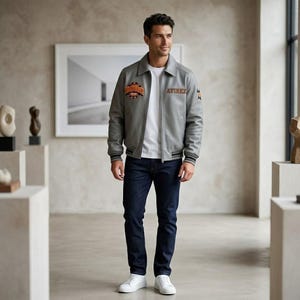 May include: A light gray leather bomber jacket with a collar, front zipper, and ribbed cuffs, featuring an orange and black circular patch on the chest and the text AVIREX on the left side, worn over a white t-shirt and dark blue denim jeans.