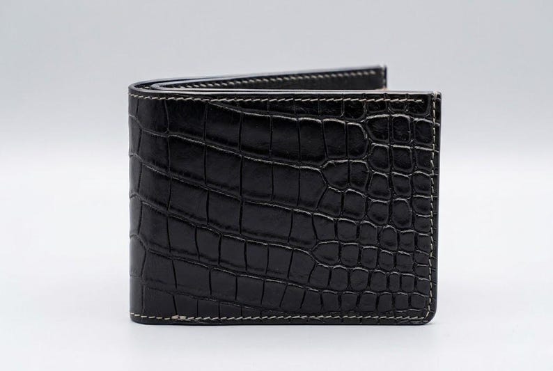 Pu&ograve; includere: Un portafoglio nero a due ante con una texture a pelle di coccodrillo. Il portafoglio &egrave; in pelle e presenta cuciture bianche sui bordi. Il portafoglio &egrave; chiuso e poggia su uno sfondo bianco.