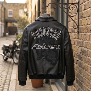 K&ouml;nnte beinhalten: Schwarze Lederjacke mit wei&szlig;em Schriftzug "TRAPSTAR" und "Avirex". Die Jacke hat ein Drachendesign auf dem R&uuml;cken. Sie h&auml;ngt an einem dekorativen Metallhaken vor einer Backsteinmauer.
