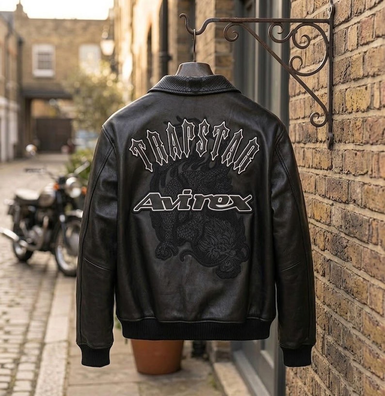 K&ouml;nnte beinhalten: Schwarze Lederjacke mit wei&szlig;em Schriftzug "TRAPSTAR" und "Avirex". Die Jacke hat ein Drachendesign auf dem R&uuml;cken. Sie h&auml;ngt an einem dekorativen Metallhaken vor einer Backsteinmauer.