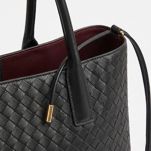 Op de afbeelding: Zwarte leren tote bag met een geweven patroon. De tas heeft twee handvatten en een schouderriem. De binnenkant is gevoerd met een bordeauxrode stof. Een goudkleurig metalen detail is zichtbaar op de riem.