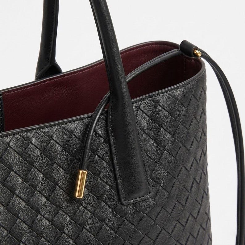 Op de afbeelding: Zwarte leren tote bag met een geweven patroon. De tas heeft twee handvatten en een schouderriem. De binnenkant is gevoerd met een bordeauxrode stof. Een goudkleurig metalen detail is zichtbaar op de riem.