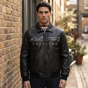 K&ouml;nnte beinhalten: Schwarze Lederjacke mit dem wei&szlig;en Text "Avi Rex TRAP STAR". Die Jacke hat einen Rei&szlig;verschluss, einen Kragen und gerippte B&uuml;ndchen und Saum. Die Person steht vor einer Backsteinmauer.