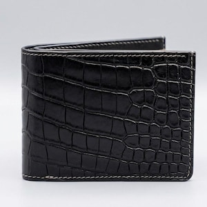 Pu&ograve; includere: Un portafoglio nero a due ante con un motivo strutturato a forma di coccodrillo. Il portafoglio &egrave; realizzato in pelle e presenta cuciture bianche sui bordi. Il portafoglio &egrave; chiuso e si trova in posizione verticale.