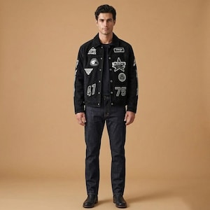 May include: A black suede jacket adorned with various white patches and the numbers "47" and "75". The jacket is worn with dark wash denim jeans and black leather boots. The model is standing against a tan backdrop.