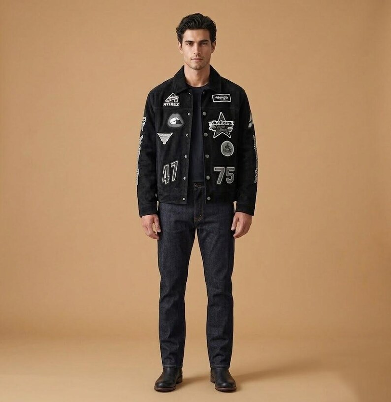 May include: A black suede jacket adorned with various white patches and the numbers "47" and "75". The jacket is worn with dark wash denim jeans and black leather boots. The model is standing against a tan backdrop.
