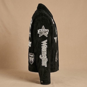 May include: Black suede jacket with white patches and the Wrangler logo. The jacket features a classic collar, buttoned cuffs, and a button closure. Patches include a star and the words "Avirex All Star Coolers".