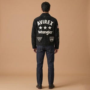 May include: A black suede jacket with white text that reads "AVIREX" above three white stars and the word "Wrangler". The jacket has patches and an American flag on the sleeve. The person is wearing dark blue jeans and black shoes.