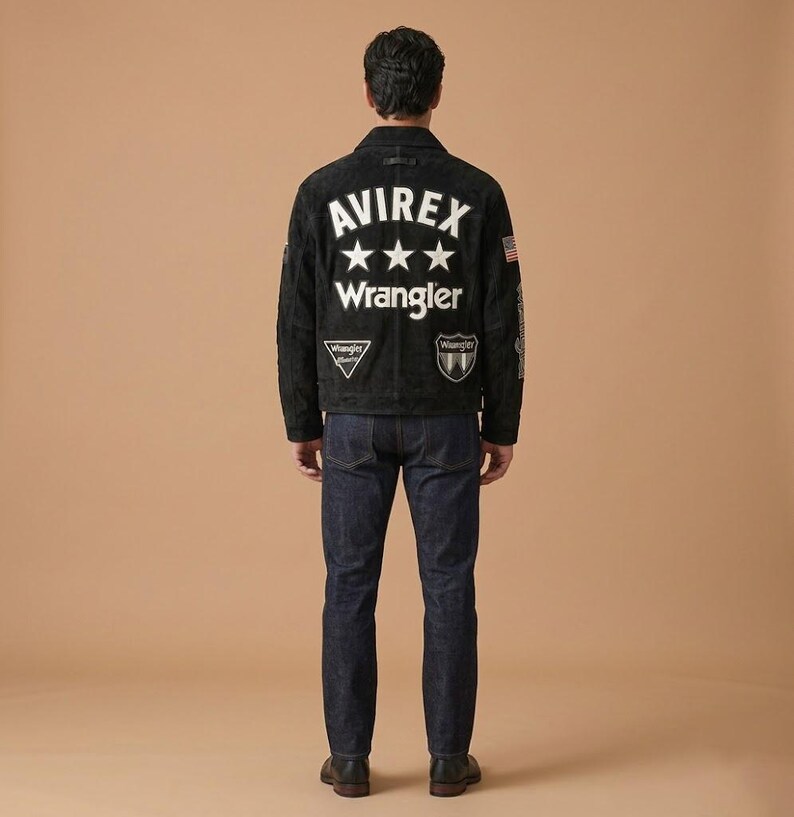 May include: A black suede jacket with white text that reads "AVIREX" above three white stars and the word "Wrangler". The jacket has patches and an American flag on the sleeve. The person is wearing dark blue jeans and black shoes.