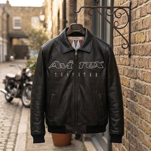 K&ouml;nnte beinhalten: Schwarze Lederjacke mit dem wei&szlig;en Text "Avi Rex Trapstar". Die Jacke hat einen Kragen, einen Rei&szlig;verschluss und schwarze gerippte B&uuml;ndchen und Saum. Sie h&auml;ngt an einem dekorativen Metallhaken vor einer Backsteinmauer.