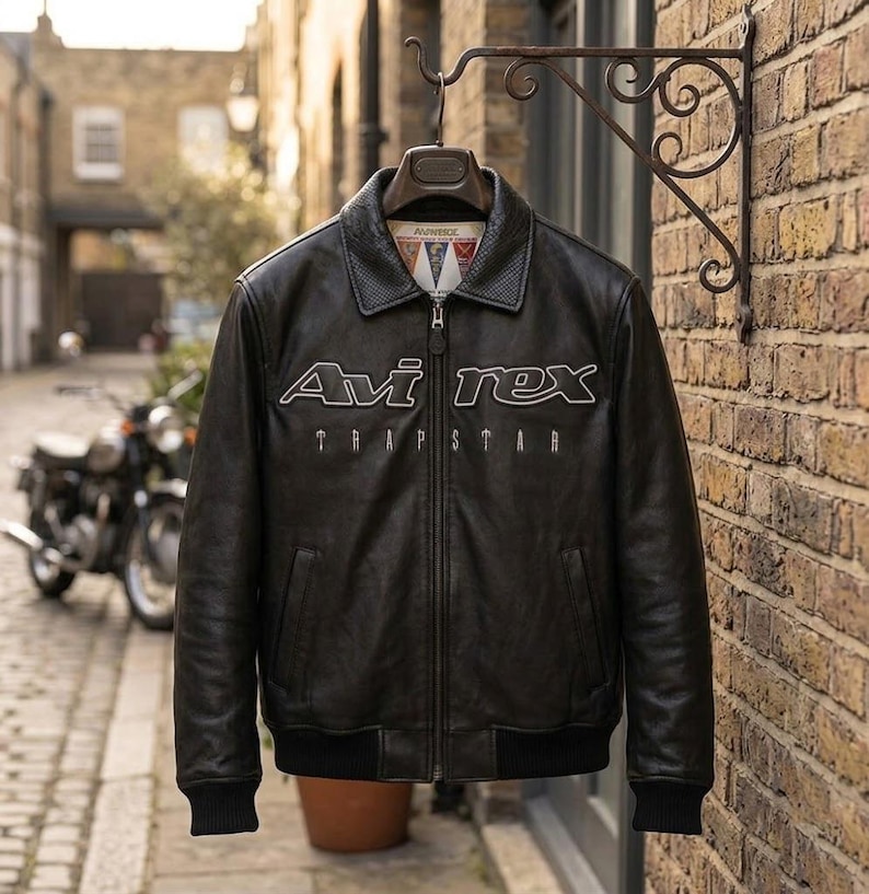 K&ouml;nnte beinhalten: Schwarze Lederjacke mit dem wei&szlig;en Text "Avi Rex Trapstar". Die Jacke hat einen Kragen, einen Rei&szlig;verschluss und schwarze gerippte B&uuml;ndchen und Saum. Sie h&auml;ngt an einem dekorativen Metallhaken vor einer Backsteinmauer.