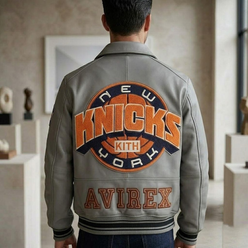 May include: A light gray leather varsity jacket featuring a large circular patch on the back with the orange and navy text NEW YORK KNICKS and KITH. Below the patch, the word AVIREX is embroidered in orange block letters with navy outlines.