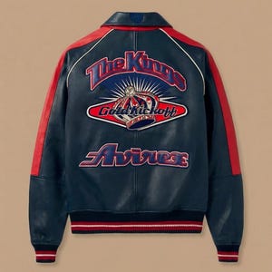 Könnte beinhalten: Marineblaue Lederjacke mit roten Akzenten an Schultern und Ärmeln. Auf dem Rücken befindet sich ein gesticktes Design mit dem Schriftzug "The Kings" über einer Krone und dem Wort "Avirex". Die Jacke hat einen gerippten Saum und Bündchen.