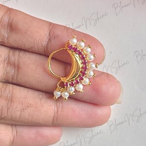 Puede incluir: Un anillo de nariz dorado con forma de media luna, adornado con una hilera de pequeñas perlas blancas en el borde exterior y una línea curva de piedras rosas vibrantes dentro de la media luna, creando una pieza de joyería detallada.