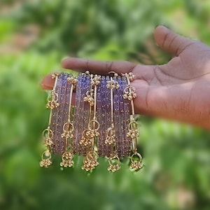 Könnte beinhalten: Eine Hand hält eine Reihe von lila Armreifen mit goldfarbenen Akzenten. Jeder Armreif hat eine Reihe kleiner, hängender goldfarbener Glöckchen. Der Hintergrund ist ein weiches, verschwommenes Grün.