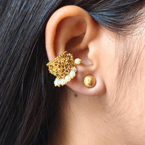 May include: A gold-toned ear cuff featuring an intricate filigree pattern with small dangling white beads, paired with a matching gold-toned stud earring. The ear cuff sits on the upper cartilage, while the stud is placed in the earlobe.
