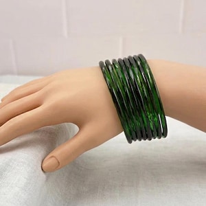 May include: A collection of dark green bangles stacked on a hand. The bangles are made of a translucent material, allowing light to pass through. The hand is resting on a white surface, and the background is a plain white wall.