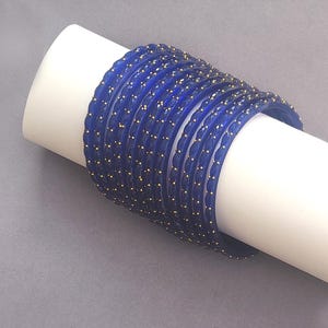 Op de afbeelding: Een stapel donkerblauwe armbanden met goudkleurige accenten. De armbanden worden gepresenteerd op een wit cilindrisch object tegen een grijze achtergrond. De armbanden hebben een textuur en zijn waarschijnlijk gemaakt van glas of een vergelijkbaar materiaal.
