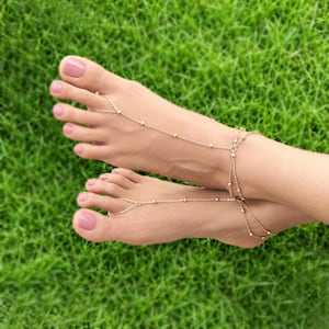 May include: A gold-toned chain foot accessory featuring delicate links and small spherical beads. The jewellery connects from a loop around the second toe, extending across the top of the foot to wrap around the ankle with a multi-strand design.