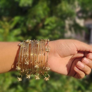 Puede incluir: Un conjunto de varias pulseras rígidas translúcidas de color marrón claro. Cada pulsera presenta finos detalles en tono dorado metálico y pequeños dijes esféricos de color dorado que cuelgan de las bandas circulares, creando un efecto.