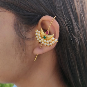 May include: A gold-toned ear cuff featuring a crescent-shaped design adorned with small white pearls. The center of the crescent is set with a small green stone, while a teardrop-shaped red stone sits above it. A thin gold wire extends from the piece.