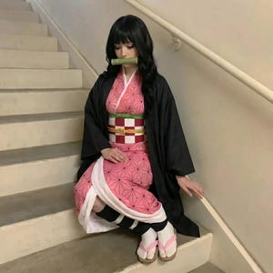 Puede incluir: Un atuendo de cosplay con un kimono rosa con un patr&oacute;n geom&eacute;trico, un abrigo negro y un obi estampado. El atuendo incluye calentadores de piernas en blanco y negro, calcetines blancos y sandalias rosas. Una boquilla de bamb&uacute; est&aacute; en su lugar.