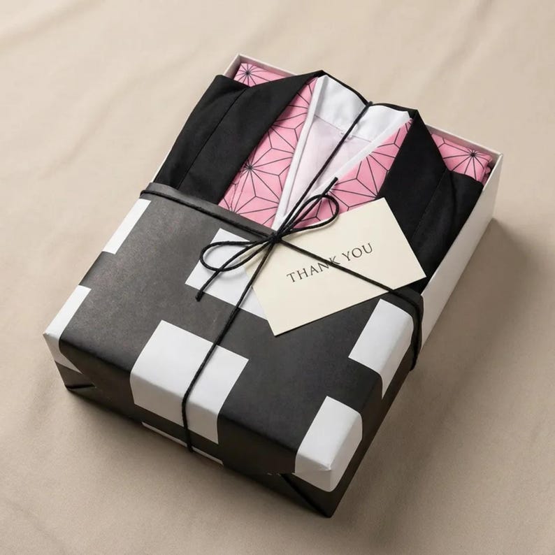 Puede incluir: Una caja de regalo envuelta en papel estampado en blanco y negro, atada con una cuerda negra y un lazo. En el interior, se ve una prenda negra y rosa. Una peque&ntilde;a tarjeta con "THANK YOU" est&aacute; adjunta.