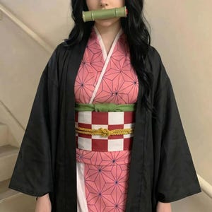 Puede incluir: Un kimono rosa con un patr&oacute;n geom&eacute;trico, un cuello blanco y un obi a cuadros rojos y blancos. El atuendo incluye un haori negro y un bozal de bamb&uacute; verde. Este atuendo de cosplay est&aacute; inspirado en un personaje de anime popular.