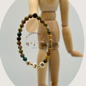 Puede incluir: Una pulsera de cuentas marrones, verdes y doradas. La pulsera incluye cuentas blancas con los números 4, 7 y 8. La pulsera se muestra frente a un maniquí de madera.
