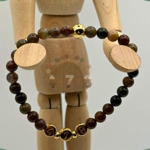 May include: A beaded bracelet with brown, green, and gold beads. The bracelet has gold accents, including a large gold bead and three smaller beads with the numbers 4, 7, and 8. The bracelet is displayed on a wooden mannequin.