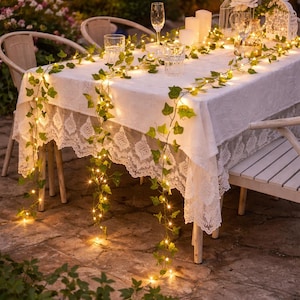May include: A garden table is set with a white lace-trimmed tablecloth, adorned with ivy garlands and fairy lights. Champagne flutes and glasses are arranged, alongside candles and a decorative birdcage, creating an inviting atmosphere for an outdoor event.