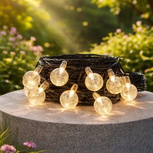 May include: A set of illuminated globe lights with a warm glow. The clear, crackled glass globes emit a soft light. The lights are arranged near a dark, woven basket, suitable for outdoor decoration. The lights are on a stone surface.