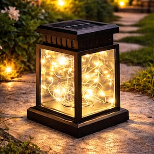 May include: Black solar-powered lantern with a rectangular shape and clear glass panels. The lantern is filled with warm white fairy lights. The top of the lantern has a solar panel. The lantern is sitting on a stone surface.
