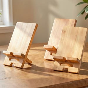 May include: Three light-coloured bamboo phone stands are arranged on a wooden table. Each stand features a rectangular back and a small ledge for holding a phone. The stands are angled for viewing.