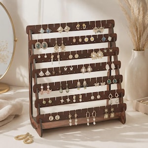 May include: A dark brown wooden earring display stand with six horizontal tiers featuring evenly spaced notches for hanging various styles of earrings, including studs, hoops, and dangling designs, arranged in a tiered, open-frame structure.