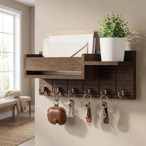 May include: A dark brown wooden wall-mounted organizer featuring a mail slot, a small shelf holding a white potted plant, and six bronze-toned metal hooks. Several sets of keys and a brown leather case are hanging from the hooks, with envelopes.