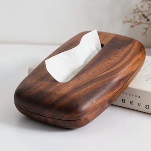 May include: A wooden tissue box with a rich, dark brown finish. The box has a rectangular shape with rounded edges and a long, oval opening. White tissues are partially visible, emerging from the top. The box is resting on a white surface.