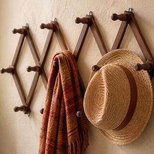 May include: An expandable dark brown wooden accordion wall rack featuring multiple pegs. A textured, multi-colored orange and red scarf hangs from one of the pegs, while a light tan straw fedora hat with a brown band rests on another peg.
