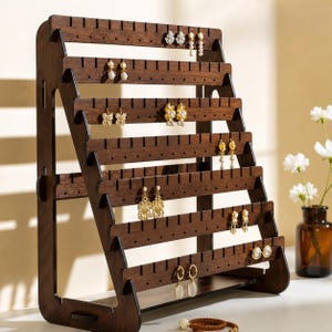 May include: A dark brown wooden earring display stand featuring six tiered horizontal shelves with pre-cut slots and holes for organizing jewelry. The structure has a sturdy side-frame design and holds various gold and pearl earrings.