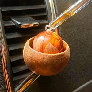 Puede incluir: Ambientador de coche de madera con un difusor esférico de color ámbar dentro de un pequeño cuenco de madera. Una pipeta de vidrio con líquido ámbar se coloca sobre el difusor. Diseño natural y minimalista.