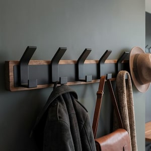 May include: A contemporary wall-mounted coat rack featuring six black metal hooks set on a wooden base. A brown hat, a brown leather bag, and a tan scarf hang from the hooks, alongside a dark jacket. The rack is on a grey wall.