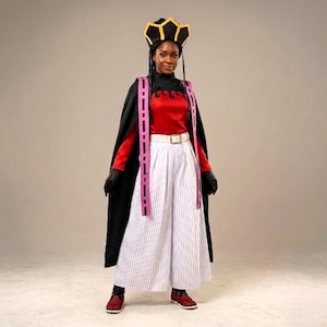 May include: A person wearing a costume with a black cape, red top, and white striped pants. The costume includes a black and gold crown, pink and black accents, and red shoes. The outfit is completed with black gloves and a white belt.