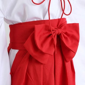 Può includere: Primo piano di un tradizionale hakama giapponese. L'immagine mostra un hakama rosso vibrante con un obi abbinato legato a un grande fiocco. L'hakama è abbinato a un top bianco, creando un contrasto sorprendente. L'outfit è probabilmente per un'arte marziale o un'occasione cerimoniale.