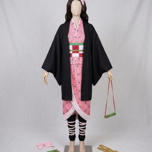 Può includere: Un costume cosplay completo con un kimono rosa a motivi geometrici, un cappotto nero e leggings a righe bianche e nere. L'outfit, esposto su un manichino, include una parrucca con un fiocco rosa e un accessorio per la museruola di bambù.