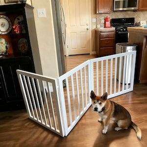 May include: A white wooden pet gate with vertical slats is positioned on a hardwood floor. A brown and white dog sits near the gate. The gate is in a kitchen setting.