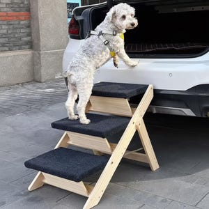 Puede incluir: Una escalera para mascotas de madera clara con tres escalones, cada uno cubierto con tela gris oscuro. Los escalones están en ángulo y conducen al maletero de un coche blanco. Un perro pequeño y blanco con arnés usa la escalera.