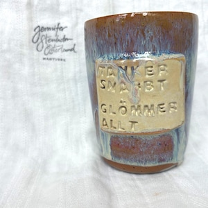 Może przedstawiać: Ręcznie wykonany ceramiczny kubek z brązowym i niebieskim szkliwem. Kubek posiada prostokątną, beżową plakietkę z wytłoczonymi słowami "TÄNKER SNABBT GLÖMMER ALLT". Kubek jest umieszczony na białym tle.