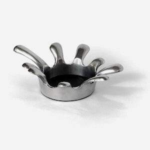 May include: A silver-colored, modern ashtray with a unique, organic design. The ashtray features a circular base with a dark interior and stylized, petal-like extensions around the rim. The overall shape resembles a stylized flower or splash.