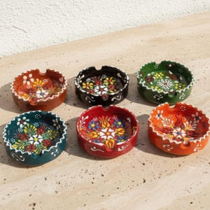 Peut inclure: Six cendriers en céramique colorés avec des motifs floraux. Les cendriers ronds ont une petite encoche pour tenir les cigarettes. Les couleurs comprennent le rouge, l'orange, le vert, le noir et le bleu sarcelle, chacun avec un motif floral unique.